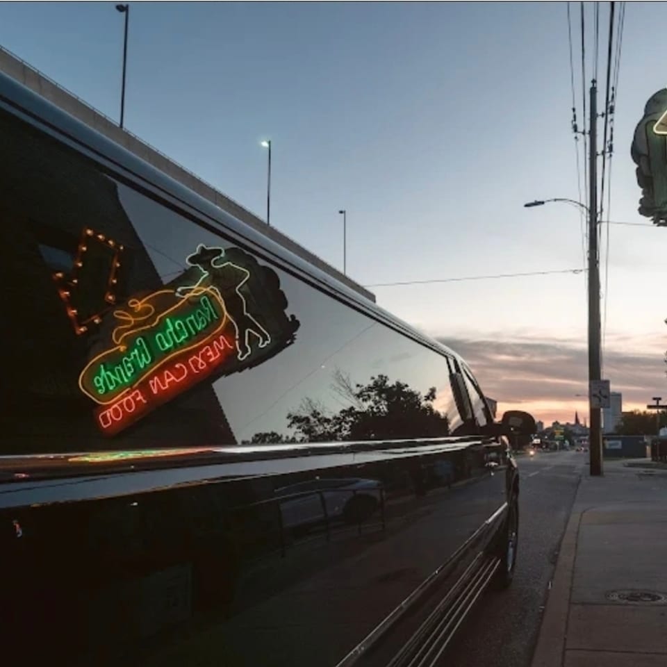 Black vehicle reflects a colorful neon restaurant sign at dusk on a sidewalk beneath an overpass.