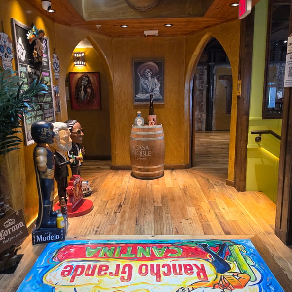 Colorful Mexican restaurant entrance with wooden flooring, tequila barrel, skeleton paintings, mannequins, chalkboard menu, and festive mural rug.