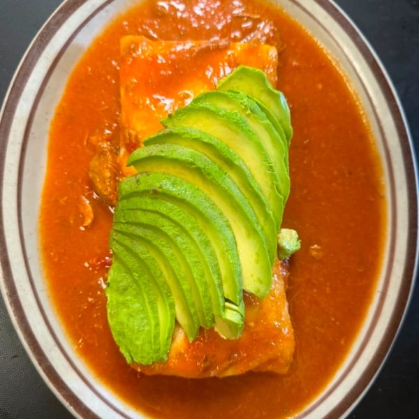 A dish with a red sauce, topped with sliced avocado, served in a brown and white striped bowl.