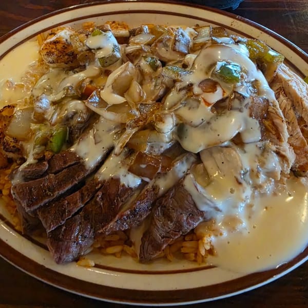 A plate of steak, chicken, and shrimp covered in white cheese sauce, onions, and peppers.
