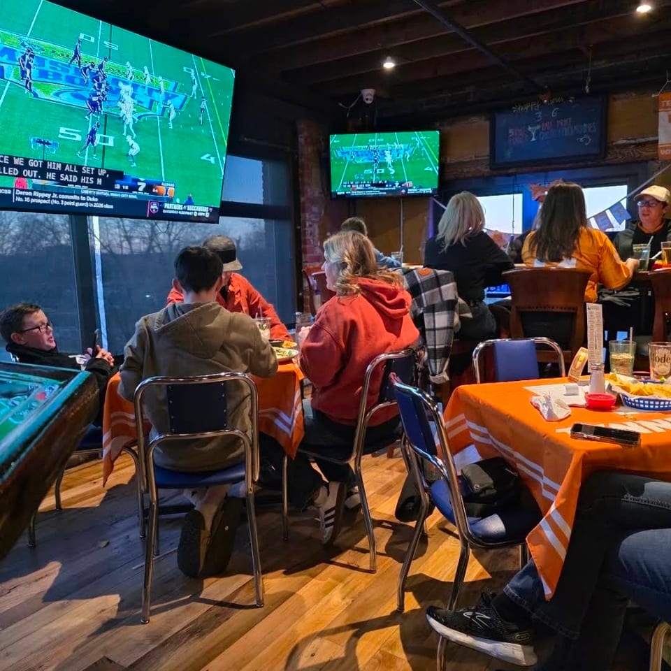 Lively sports bar with diners, orange tablecloths, food, and multiple televisions showing a football game.