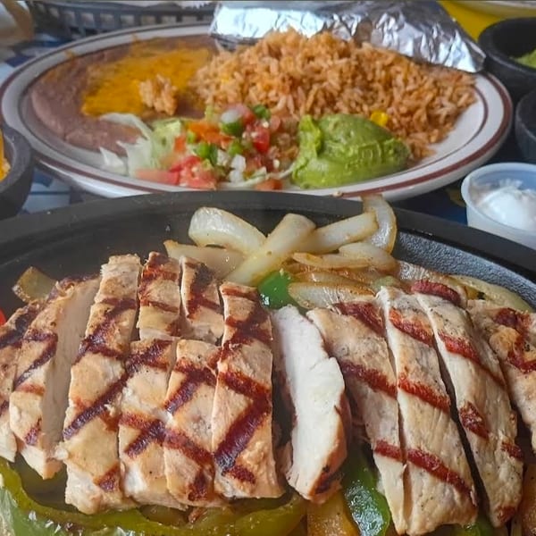 Grilled chicken fajitas with onions and peppers, served alongside Mexican rice, beans, guacamole, pico de gallo, cheese, and sour cream.