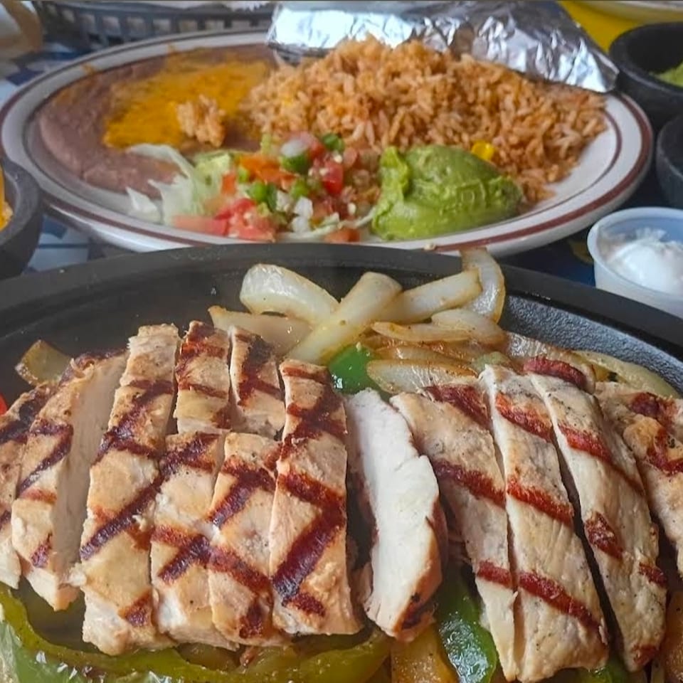 Grilled chicken fajitas with onions and peppers, served alongside Mexican rice, beans, guacamole, pico de gallo, cheese, and sour cream.