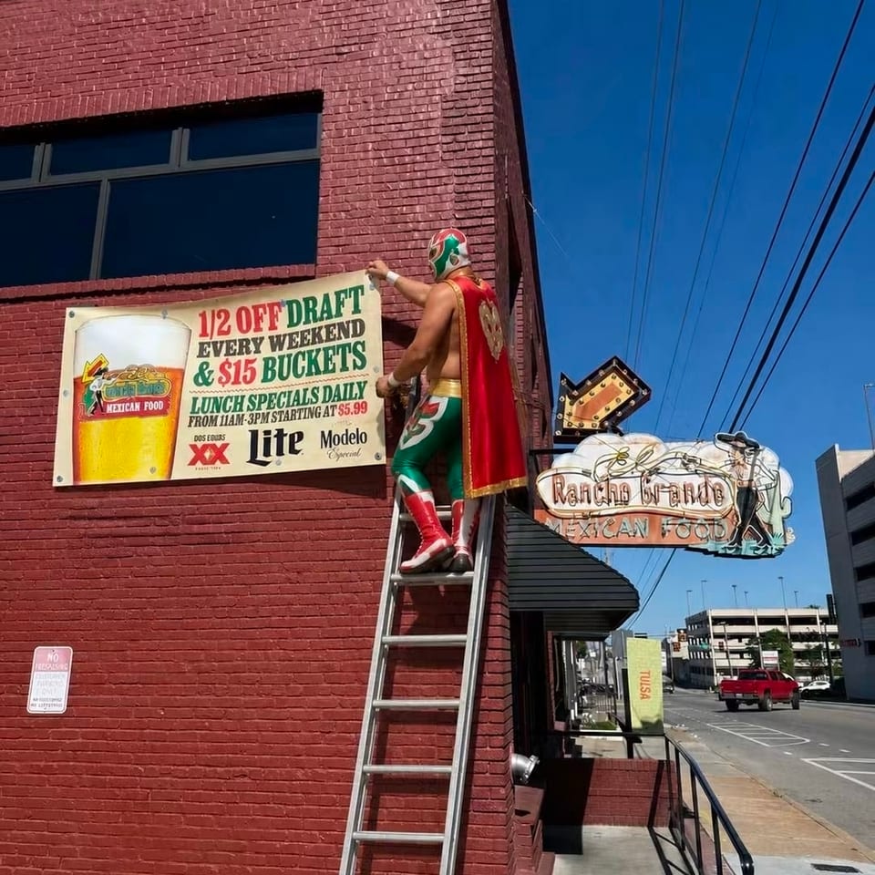 Masked luchador statue stands on a ladder beside a Mexican restaurant exterior with promotional beer signage and street view.