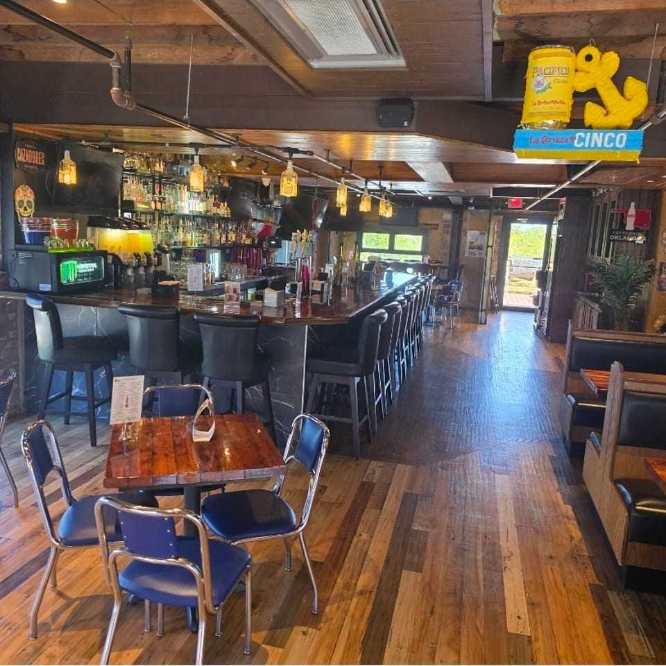 Rustic restaurant interior with a fully stocked bar, counter seating, wooden tables, booths, and colorful hanging decorations.