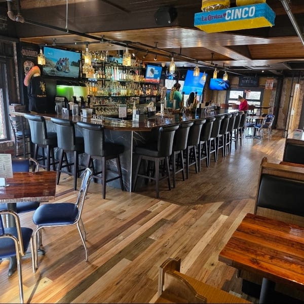 Rustic restaurant interior with a fully stocked bar, leather stools, wooden tables, booths, televisions, and large windows.