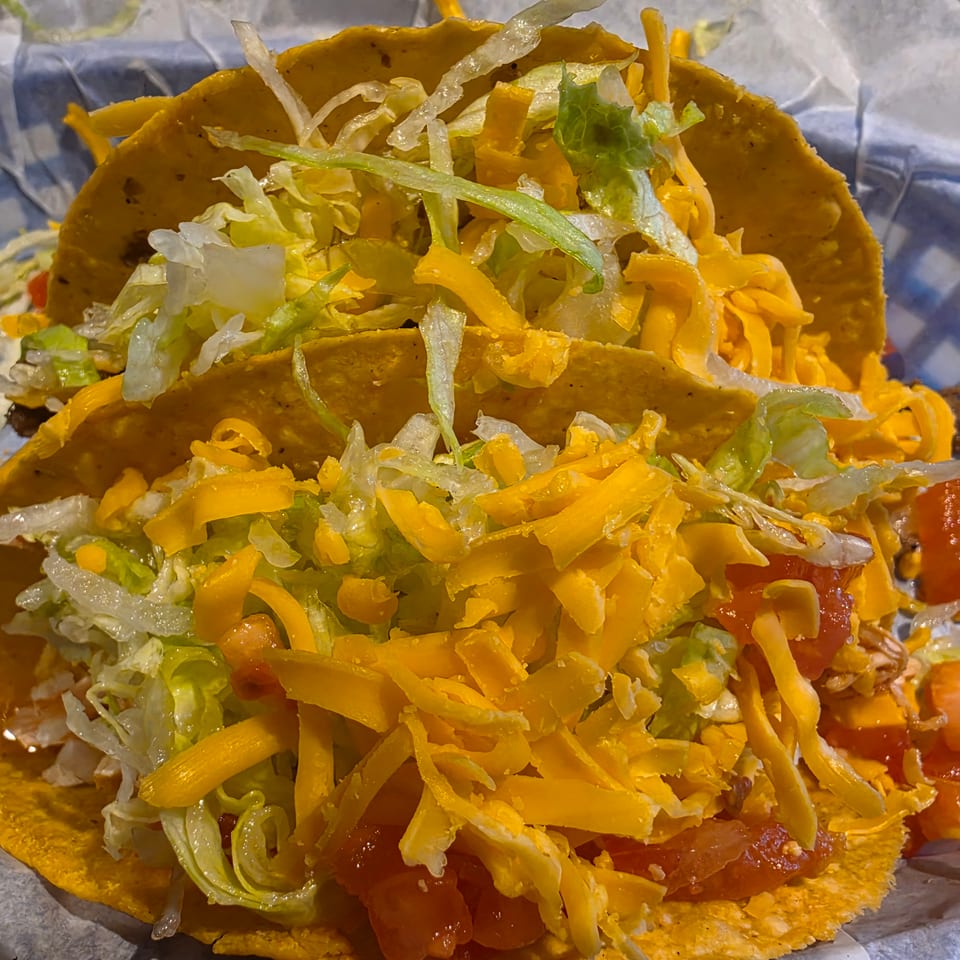 Three hard-shell tacos filled with lettuce, shredded cheddar cheese, tomatoes, and seasoned meat, served on foil.