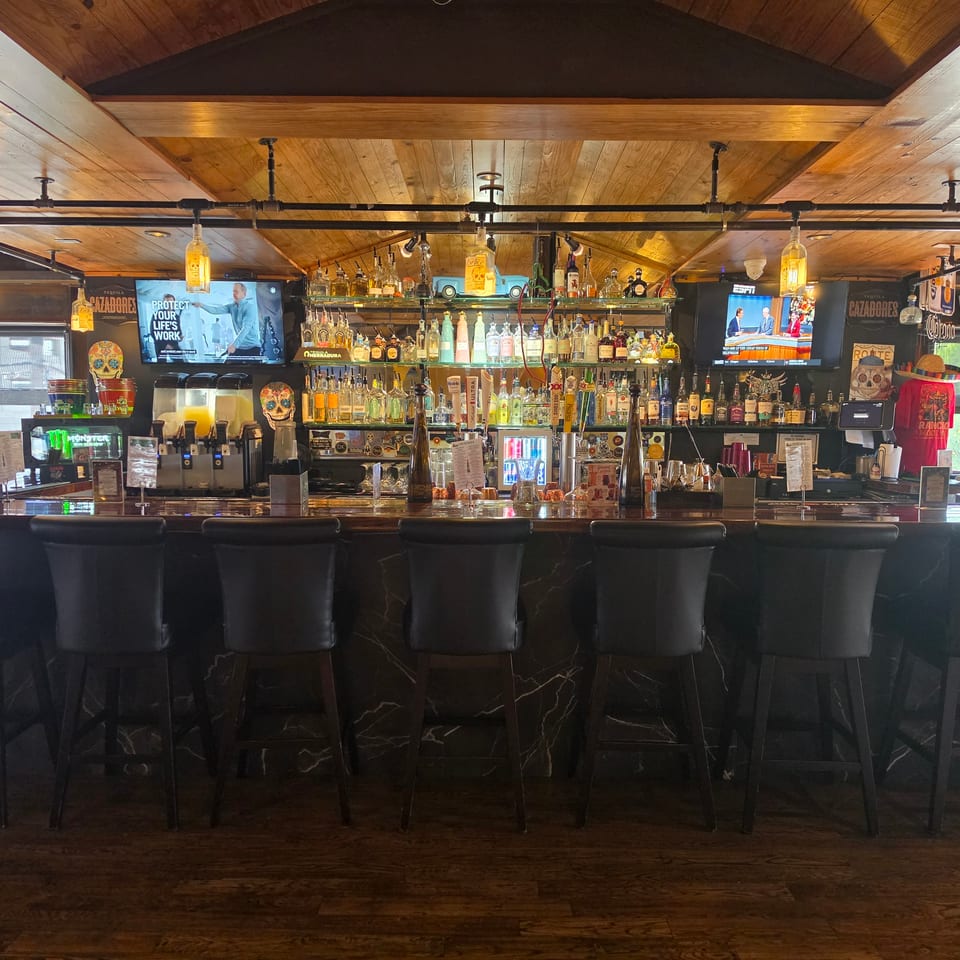 Well-stocked restaurant bar with tequila bottles, cocktail supplies, illuminated pendant lights, televisions, and rows of black barstools.