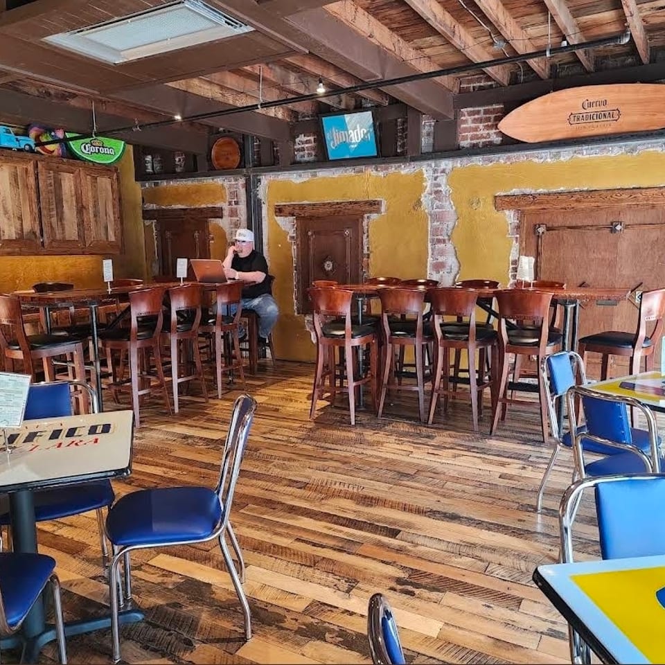 Rustic Mexican restaurant dining room with colorful tables, blue chairs, wooden floors, bar seating, and Corona decorations.