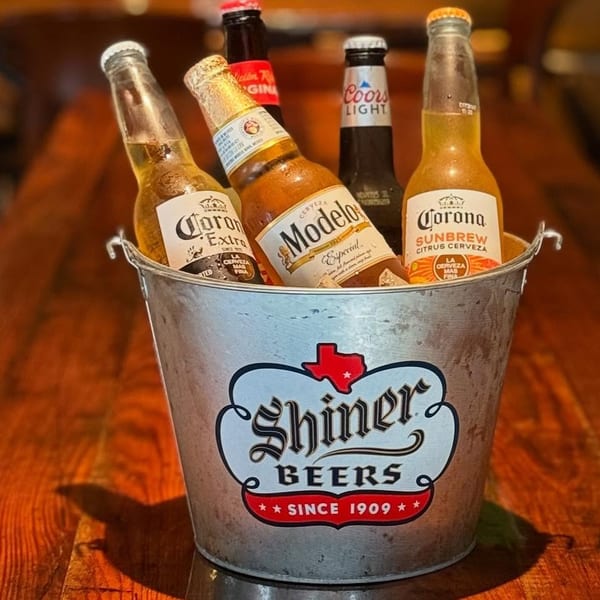 A metal Shiner Beers bucket filled with assorted beer bottles on a wooden table.