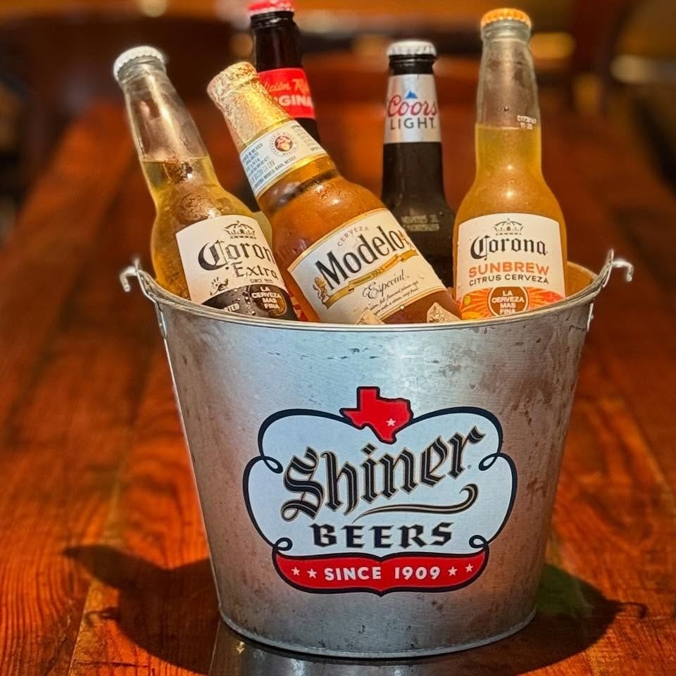 A metal Shiner Beers bucket filled with assorted beer bottles on a wooden table.