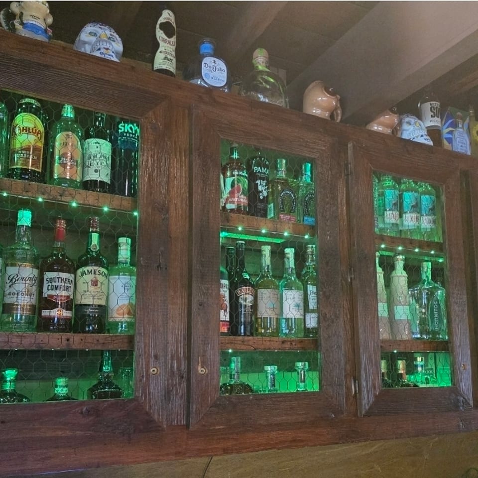Dark wooden cabinet displaying assorted liquor bottles behind green-lit wire-mesh doors in a rustic restaurant bar.