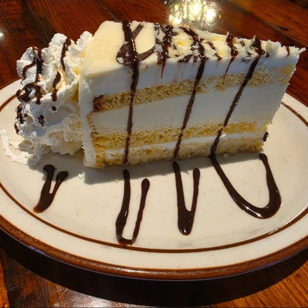 A slice of layered white cake with chocolate drizzle and whipped cream on a white plate.