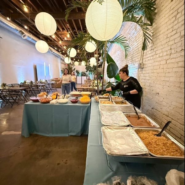 Catering buffet with Mexican food and toppings in a festive event hall with lanterns and palm decor.