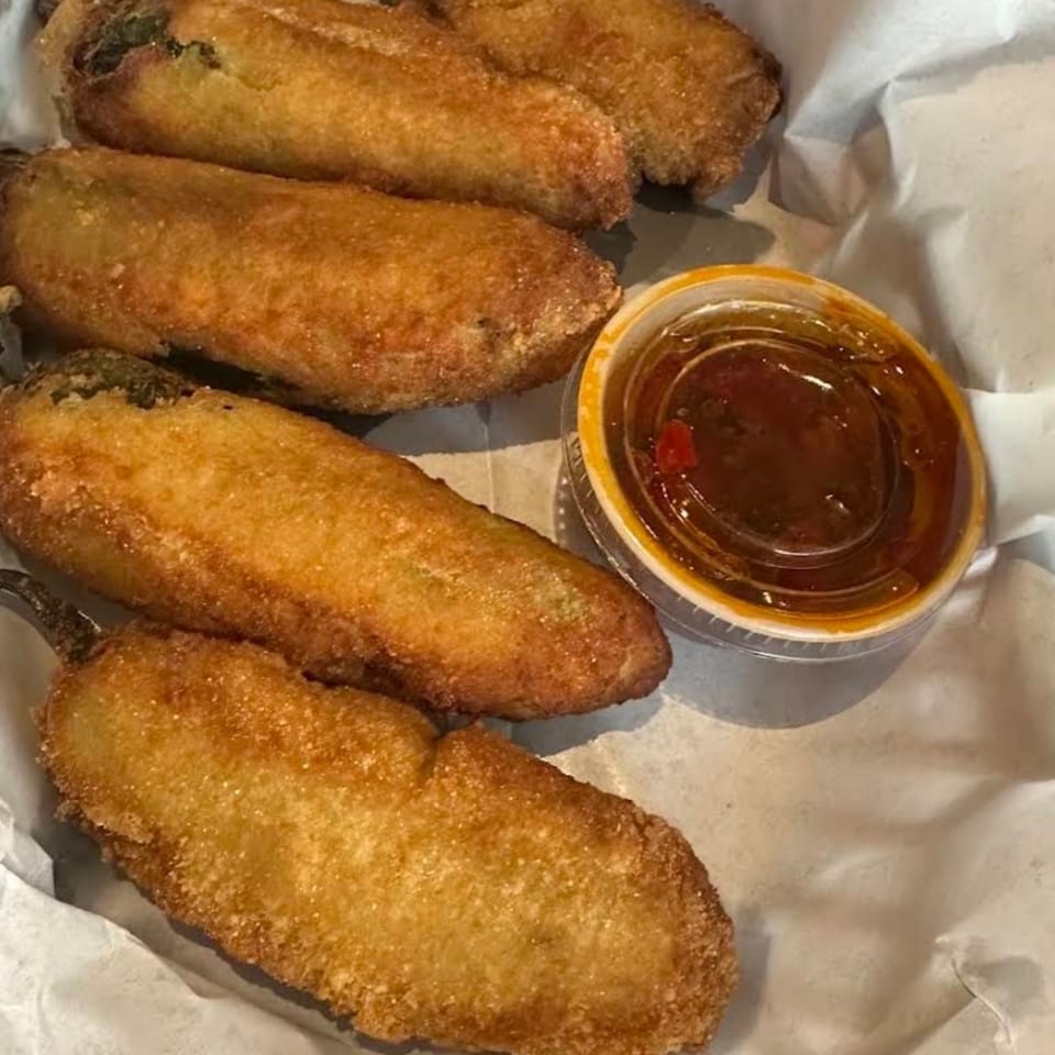 Five golden-brown fried stuffed peppers served on paper with a cup of reddish dipping sauce.