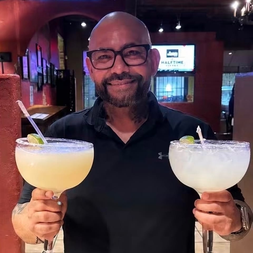 Smiling bartender holds two large lime-garnished margaritas inside a warmly lit restaurant bar.