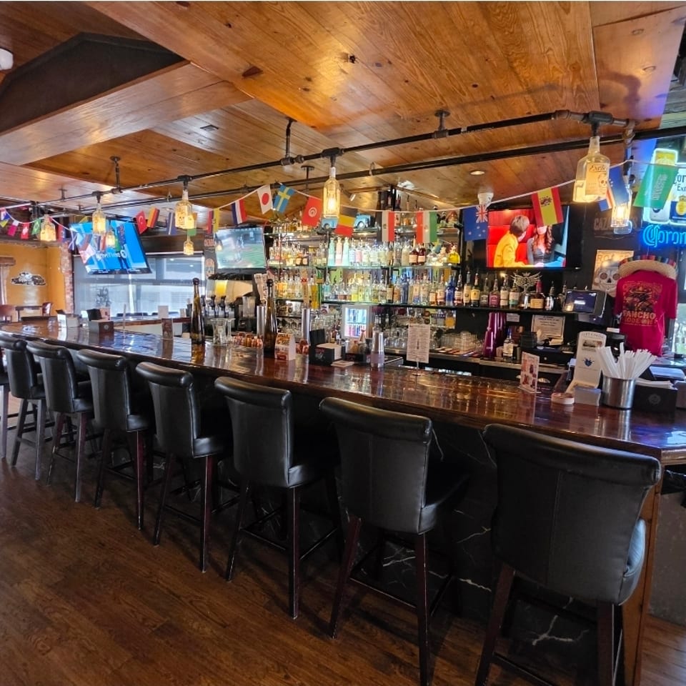 Spacious bar and restaurant interior with a long wooden counter, black stools, liquor shelves, televisions, and international flags.