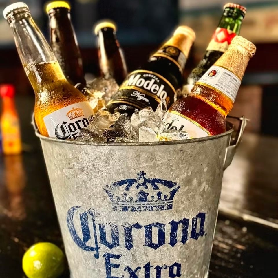 Bucket of Corona Extra and Modelo beer bottles chilling in ice, with a lime beside it on a bar.