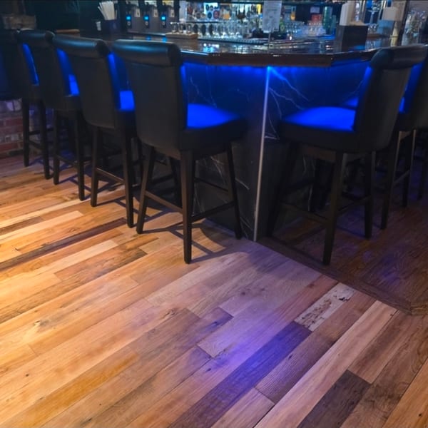 Dark restaurant bar with black leather stools, blue accent lighting, stocked shelves, and rustic wood flooring.