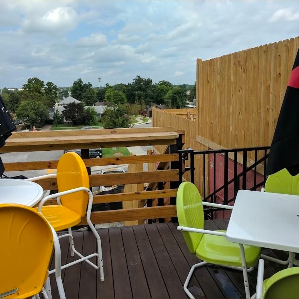 Rooftop restaurant patio with colorful chairs, white tables, black umbrellas, wooden railings, and neighborhood views.