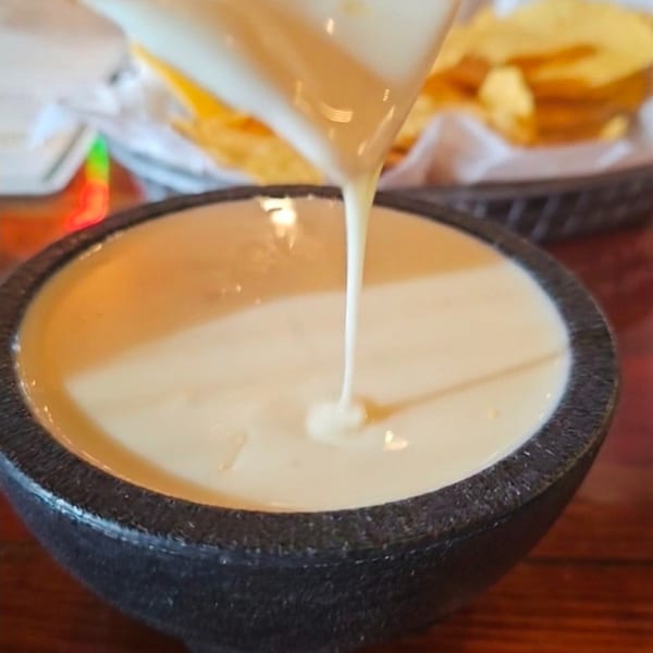 A tortilla chip dripping white queso into a black stone bowl, with more chips in the background.
