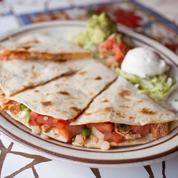 A plate of quesadillas with pico de gallo, guacamole, and sour cream on a patterned table.