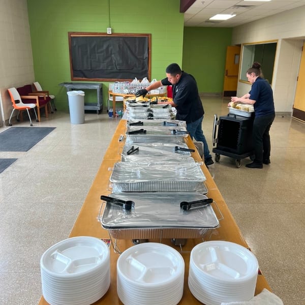 Catering staff preparing a buffet with chafing dishes and disposable plates in a room with green walls.
