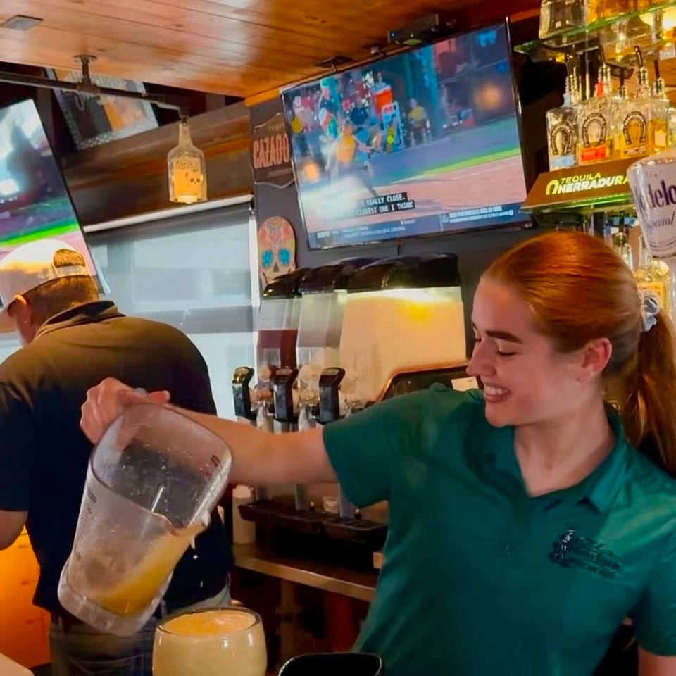 Bartender pours a frozen orange cocktail into a goblet at a warmly lit bar with tequila bottles and televisions.