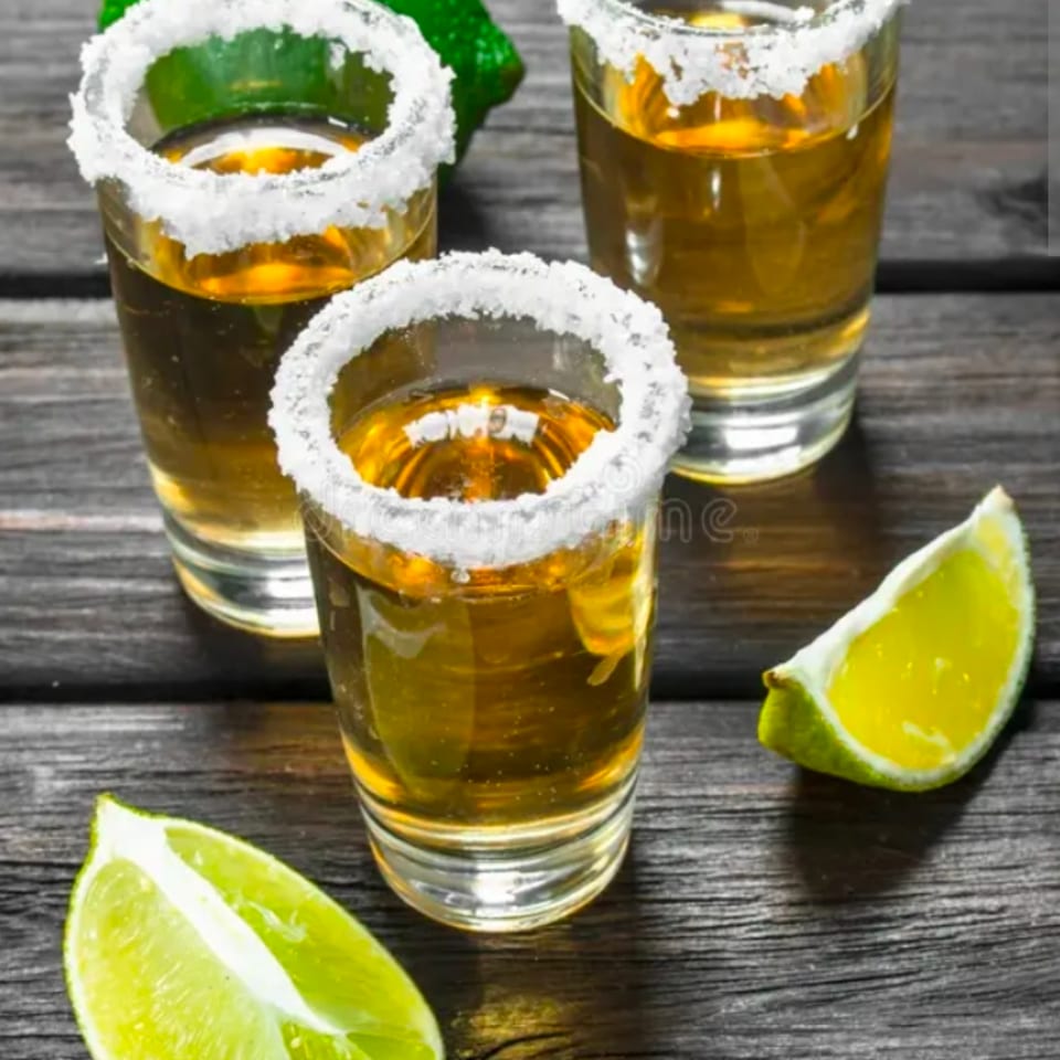 Three golden shots with salt-rimmed glasses sit beside lime wedges and a whole lime on a rustic wooden table.
