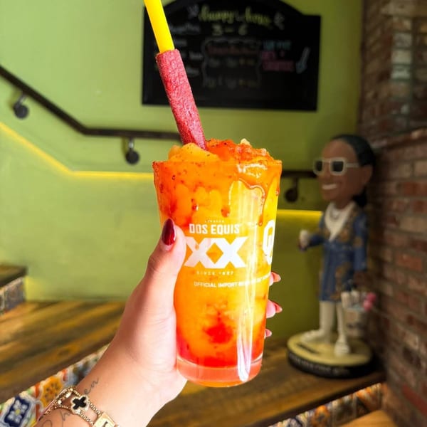Hand holding a Dos Equis glass with a vibrant orange and red frozen drink and a candy straw.