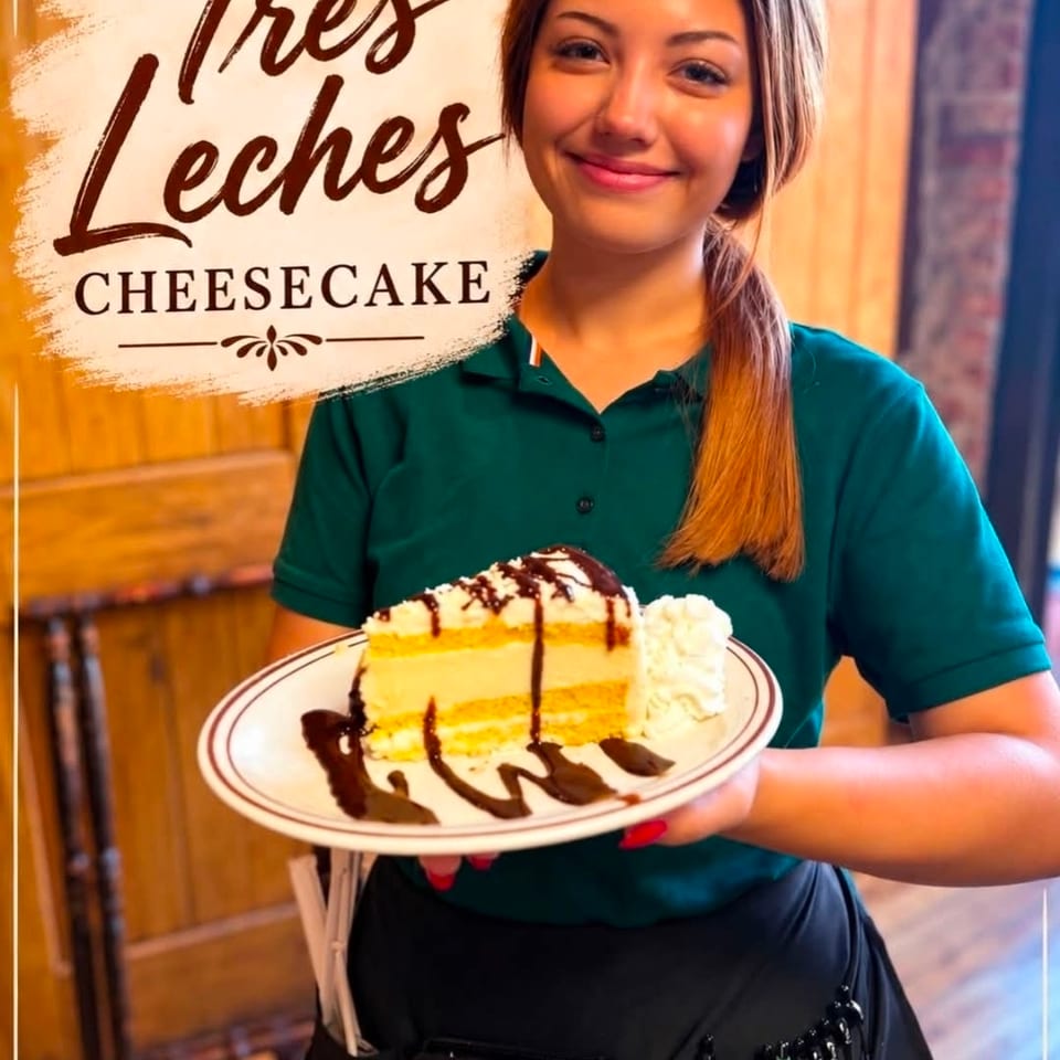 Smiling server presents a slice of chocolate-drizzled cheesecake with whipped cream at Tres Leches Cheesecake.