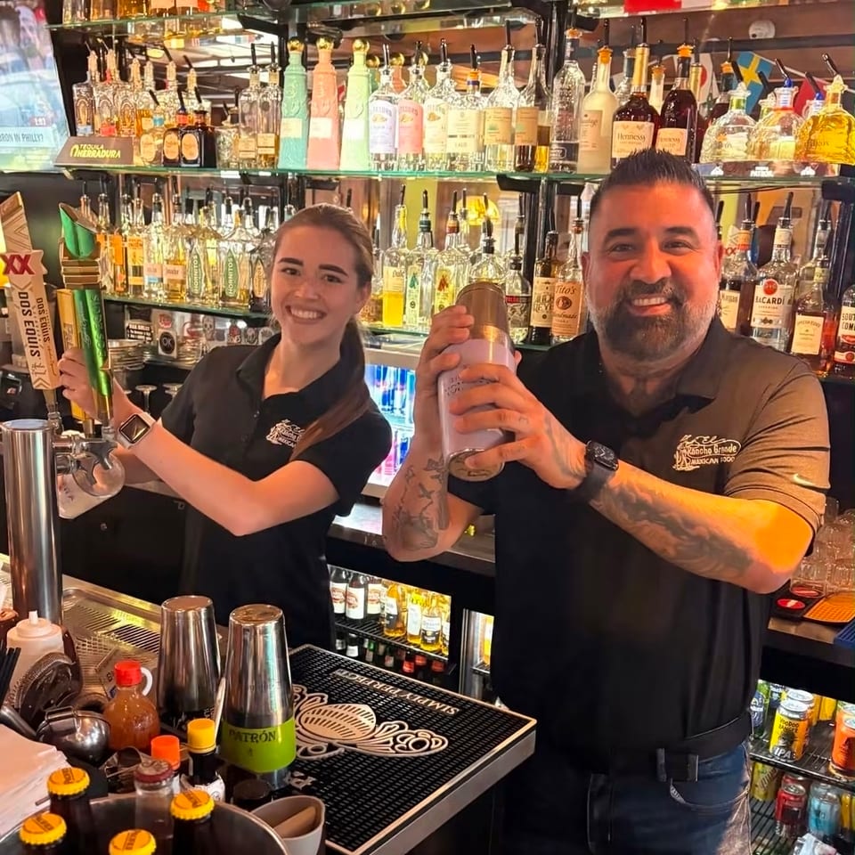 Two smiling bartenders pose behind a fully stocked bar, with one shaking a cocktail and the other holding a menu.