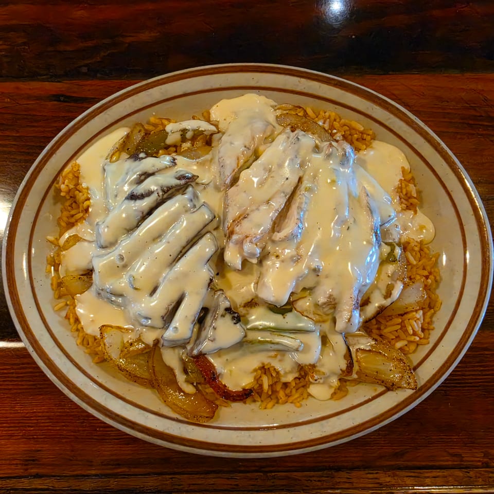Plate of grilled chicken and vegetables covered in creamy sauce, served over seasoned rice on a wooden table.
