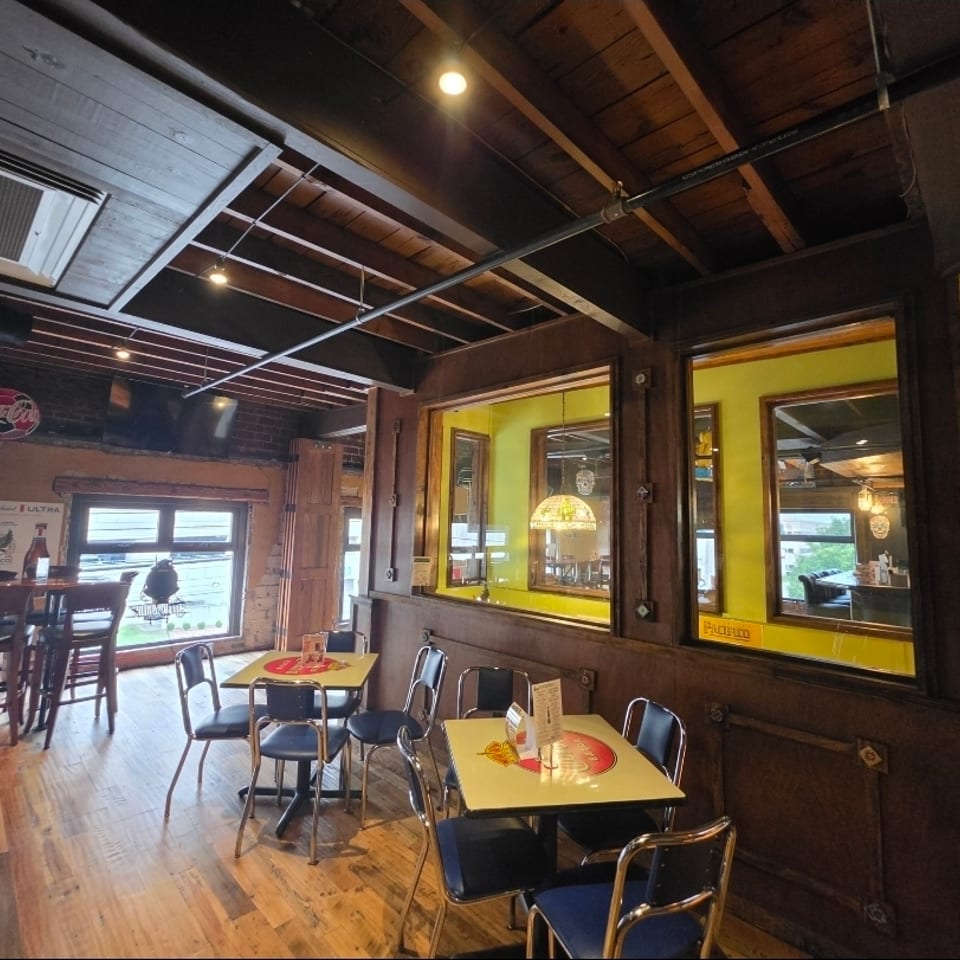 Rustic restaurant dining room with yellow tables, black chairs, exposed wooden beams, brick walls, and vintage Coca-Cola décor.