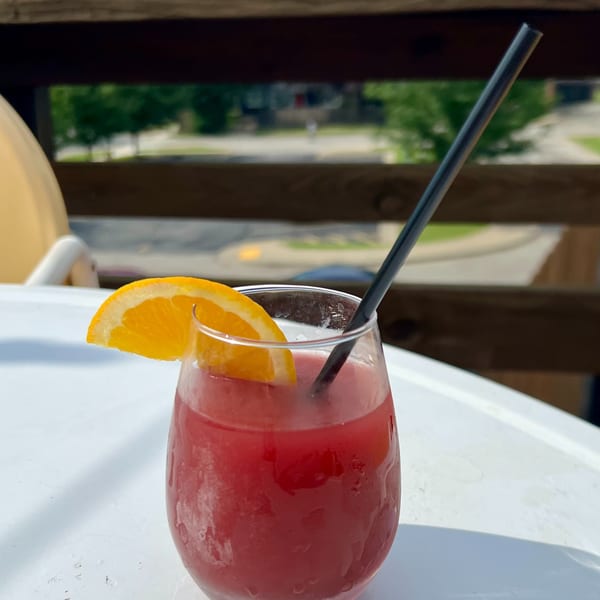 A frosty red drink with an orange slice and black straw on a white table outdoors.