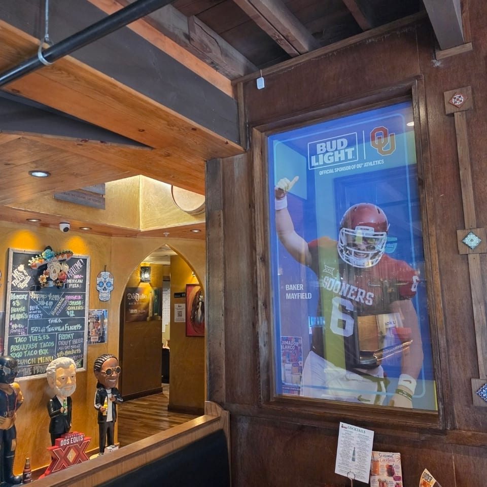 Rustic Mexican restaurant interior with wood beams, booth seating, chalkboard specials, figurines, and a Baker Mayfield football poster.