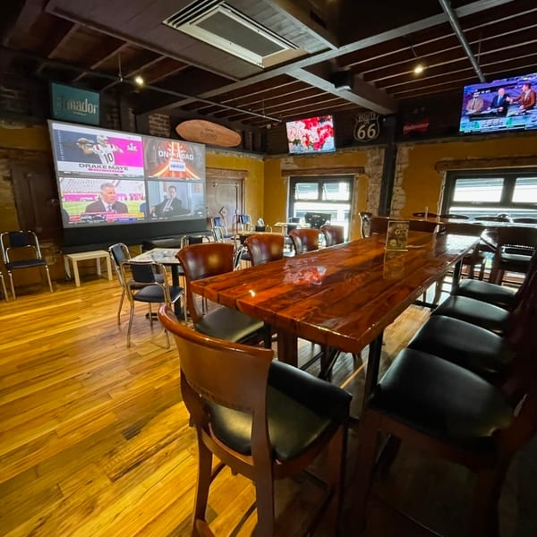 Rustic sports bar interior with a polished communal table, leather chairs, wood floors, exposed beams, and multiple televisions.