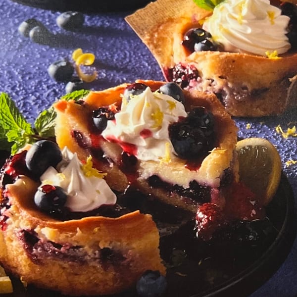 Blueberry pastries topped with whipped cream, berry sauce, fresh blueberries, lemon zest, and mint on a black plate.