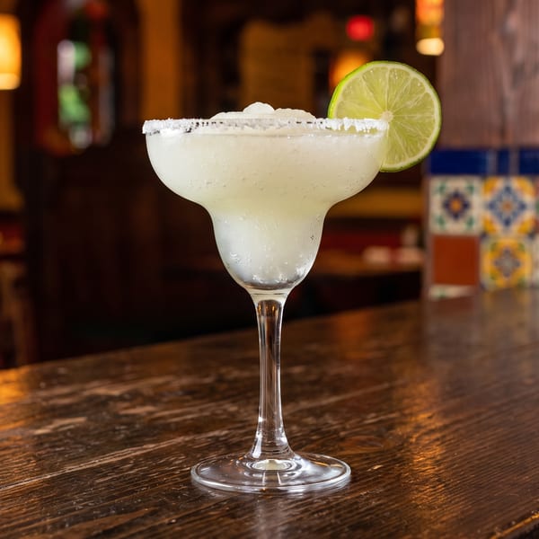 A frosty margarita with a lime slice and salt rim sits on a dark wooden bar.