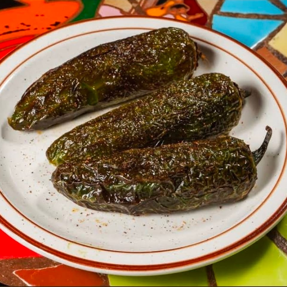 Three blistered roasted green peppers arranged on a white plate atop a colorful tiled table.