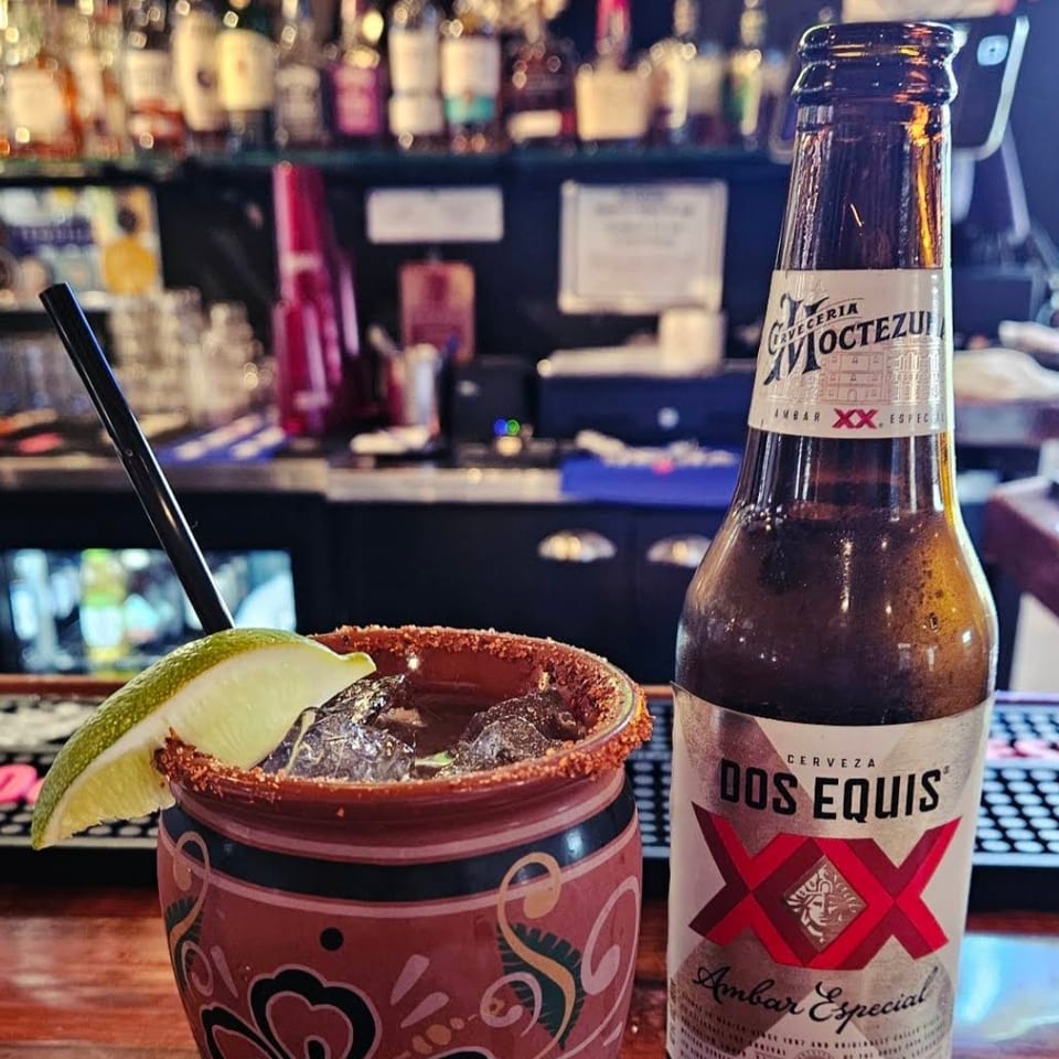 Michelada-style beer cocktail with lime wedge and chili-salted rim beside a Dos Equis beer bottle on a colorful restaurant bar.