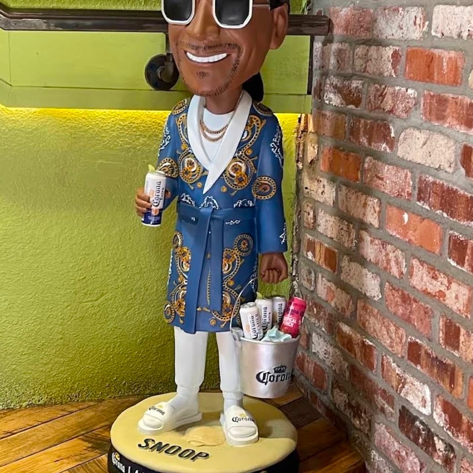 Sculpture of a smiling man in sunglasses and a patterned blue suit holding a Corona beer beside a bucket of bottles.