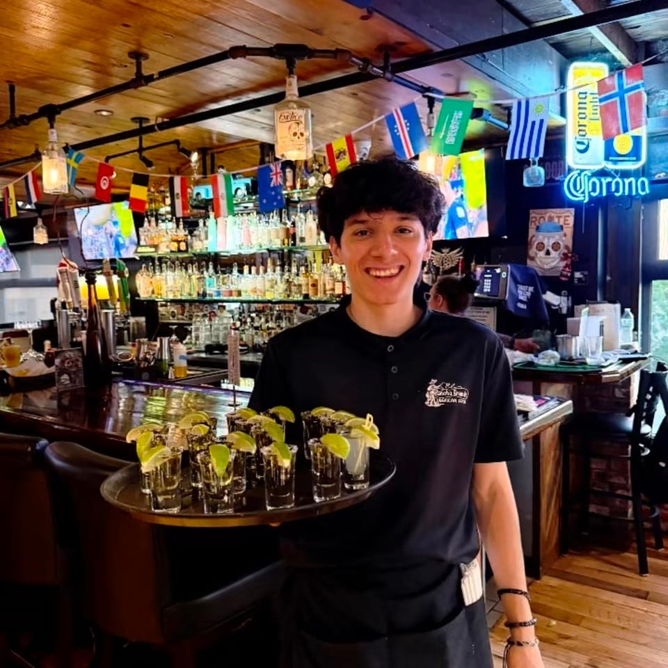 Smiling server carries a tray of lime-garnished drinks inside a warmly lit bar decorated with international flags.
