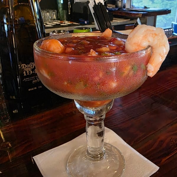 Large margarita-style glass filled with red shrimp cocktail, garnished with shrimp, on a napkin at a restaurant bar.
