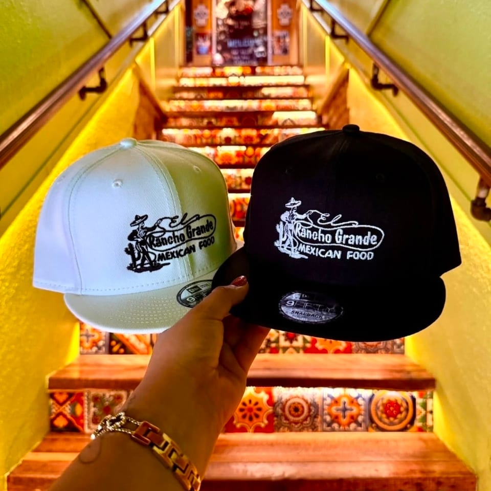 Hand holds white and black Rancho Grande Mexican Food caps over brightly tiled, warmly lit restaurant stairs.